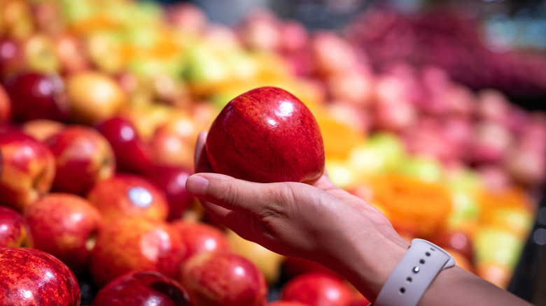 person holding shiny red apple above dozens of other apples in grocery produce department