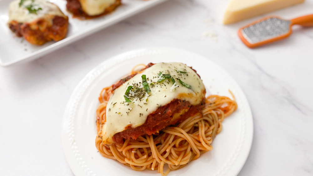 chicken Parmesan sitting on a bed of sauce and spaghetti noodles on a white plate sitting on a white and gray counter