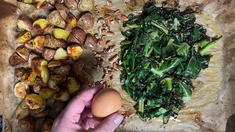 Hand cracking egg onto sheetpan with potatoes, pecans, and collard greens