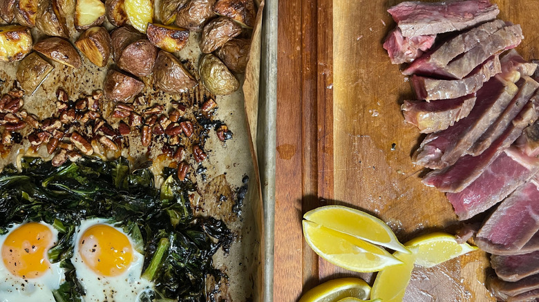 Sheet pan with potatoes, pecans, collard greens, and eggs next to cutting board with sliced steak and lemon wedges