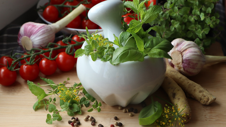 Mortar and pestle with basil surrounded by tomatoes, horseradish root, whole peppercorns