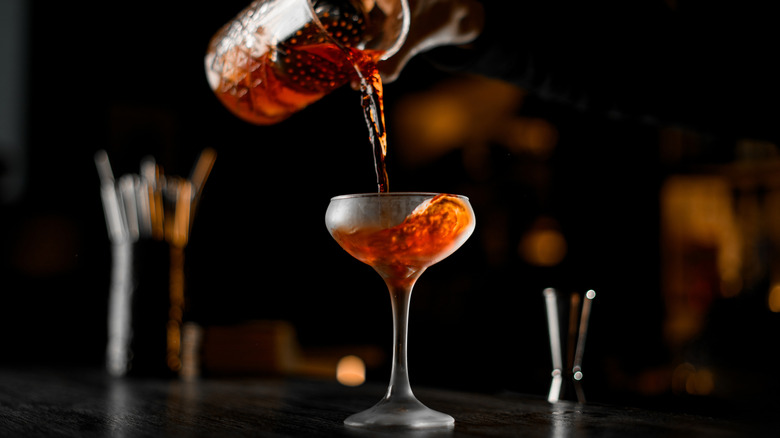 Pouring amber-colored whiskey cocktail from mixing glass into coupe glass