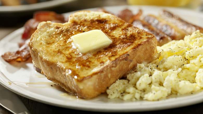 Thick French toast on a plate with butter, bacon, and scrambled eggs