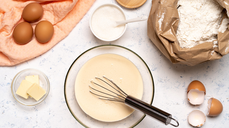 A whisk over a bowl with cake batter, with eggs, sugar, butter and flour around, over a table