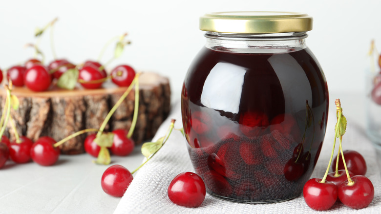 A jar of pickled cherries over a white tablecoth around more cherries