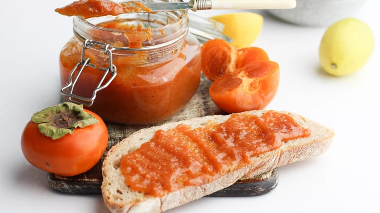 Whole and sliced persimmons with jar of jam and piece of bread.