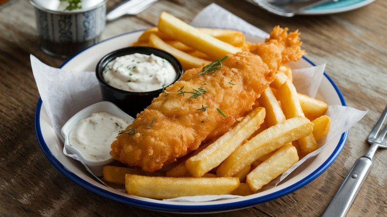 A white and blue plate of fried fish and chips with tartar sauce on a wooden table with metal utensils