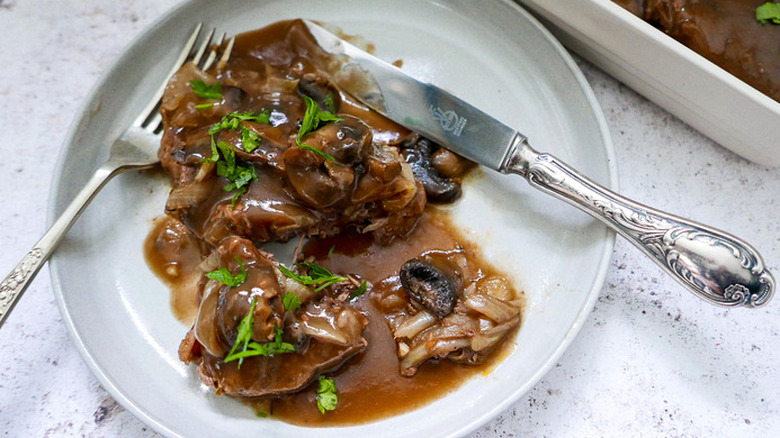 Steak, mushrooms, and gravy on white plate with utensils.