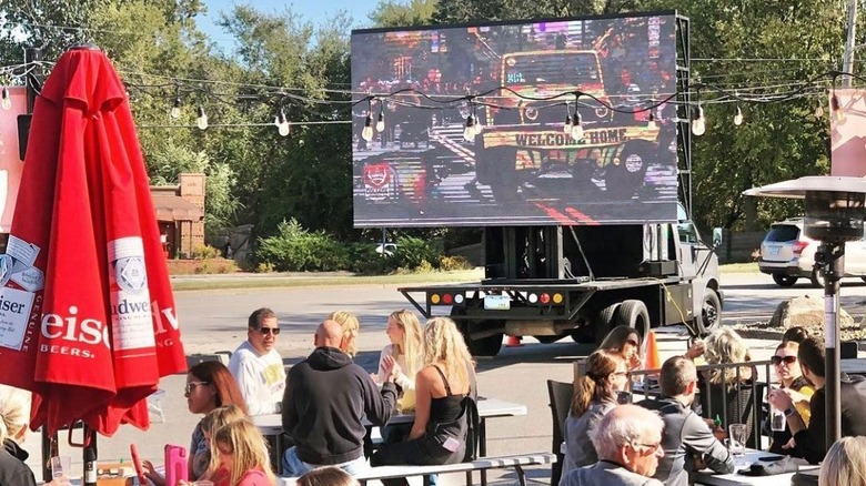 People outside at picnic tables with a TV mounted on the back of a truck. 