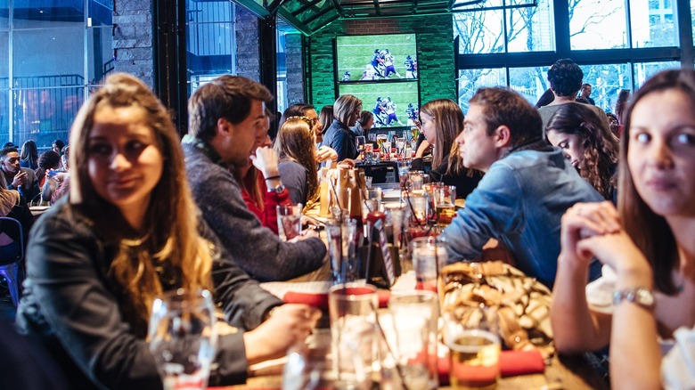 People sitting at a table in a crowded food hall with a football game playing on a TV screen in the background.