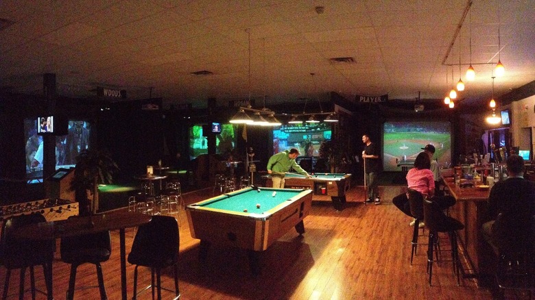 People in a dimly lit sports bar; some are playing at a pool table, while others are seated at the bar counter. 