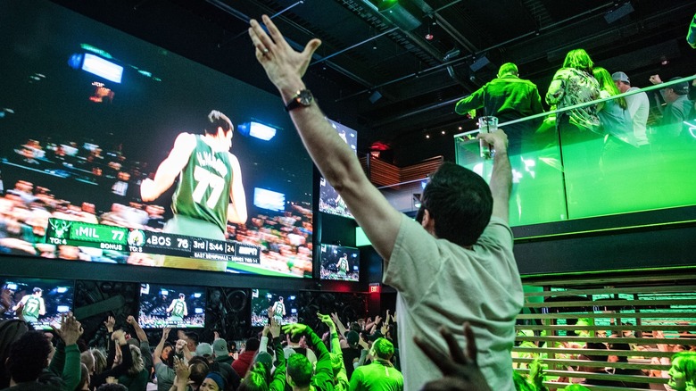 A man holding both arms with a drink in one hand, shouting at a TV screen playing a basketball game. He is in a bar packed with people, some of whom are painted in sports teams' colors. 