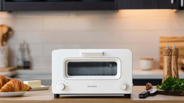 A white Balmuda toaster on a wood countertop surrounded by ingredients