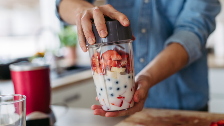 Person holding blender loaded with strawberries, blueberries, bananas, and milk in kitchen