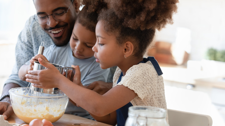 A Black man bakes with his young daughters in a bright kitchen