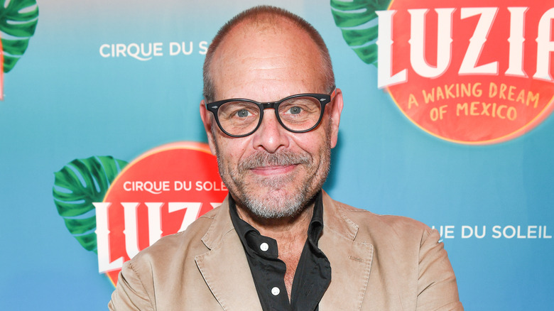 Alton Brown smiles in glasses and tan jacket