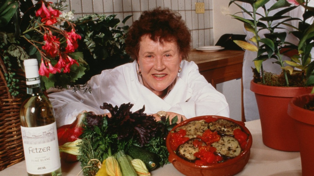 Chef Julia Child leaning on table near wine, fresh vegetables, and a cooked dish