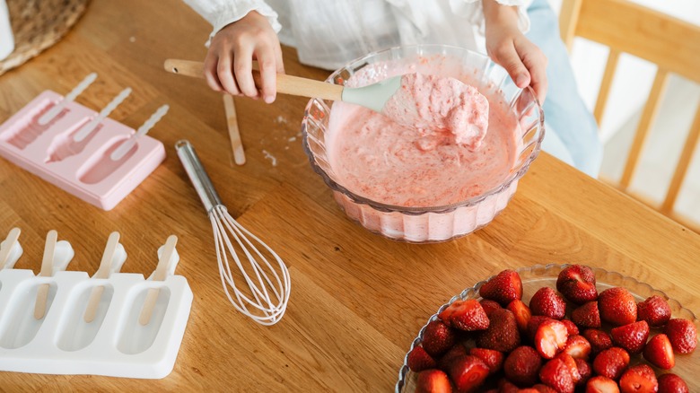 Melted strawberry ice cream in a bowl next to strawberries,whisk, and popsicle molds