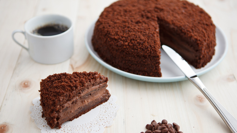 A slice of Brooklyn blackout cake with a whole cake and a cup of coffee