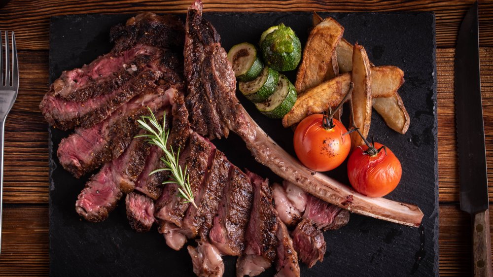 Knife, fork, and Tomahawk ribeye steak on a slate with roasted tomatoes, fries, roasted zucchini