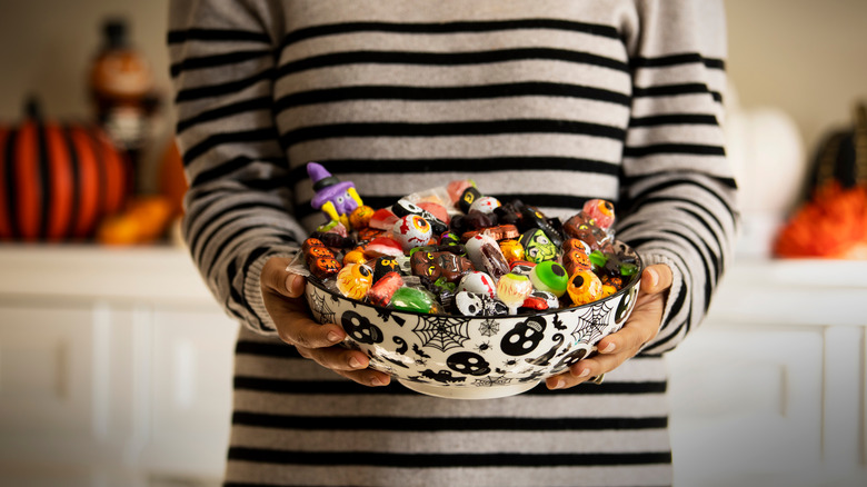 woman holding a bowl of assorted halloween candy in a halloween-themed bowl and fall decor behind her