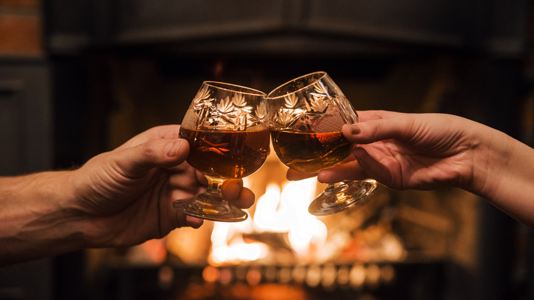 Two people making a toast with scotch by a fireplace 