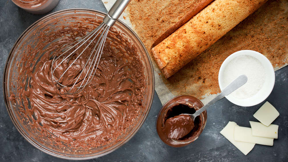 Whisk in bowl of chocolate substance, cake rolled up, small bowl of darker chocolate and white chocolate pieces