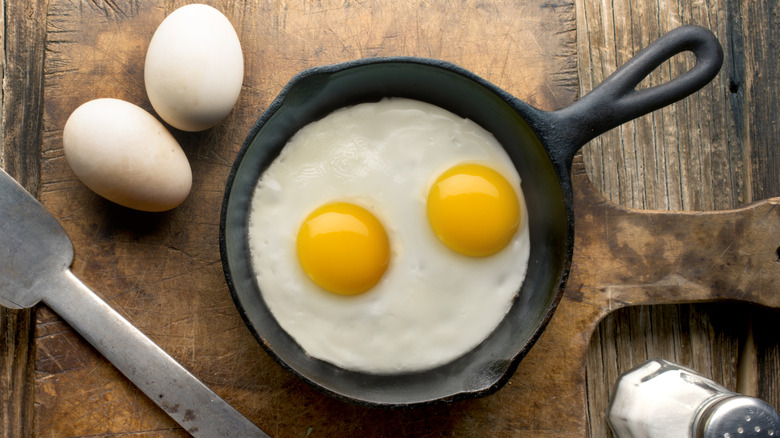 Two fried eggs in cast iron skillet next to two whole eggs