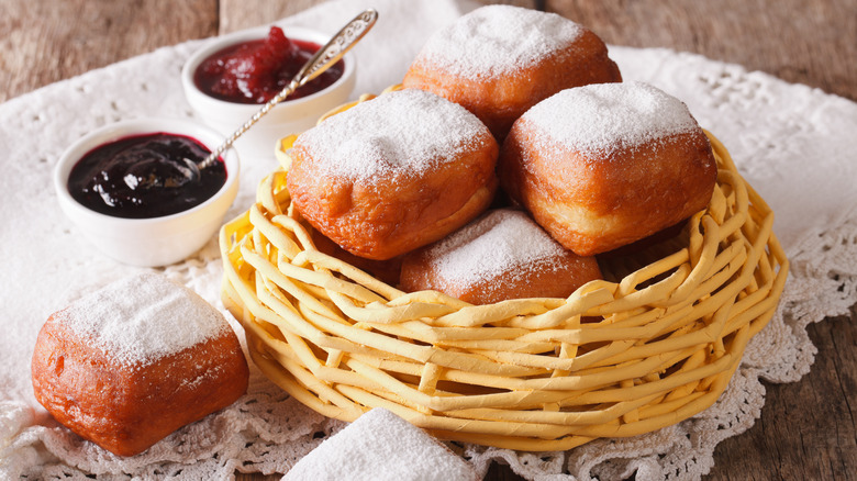 basket of beignets with powdered sugar and bowls of jam on white cotton doily on wooden table