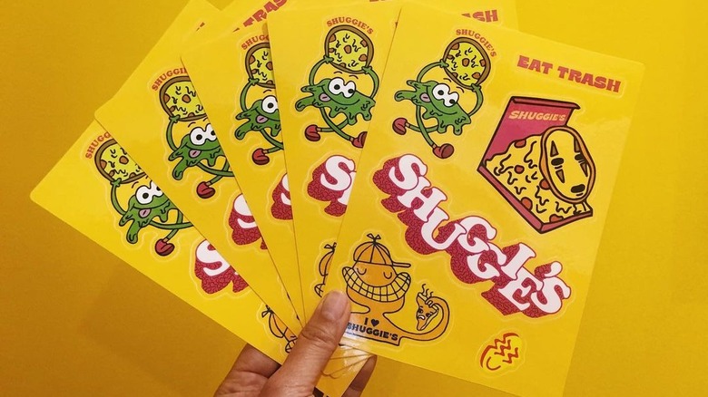 Hand holding Shuggie's pizza stickers on yellow background.