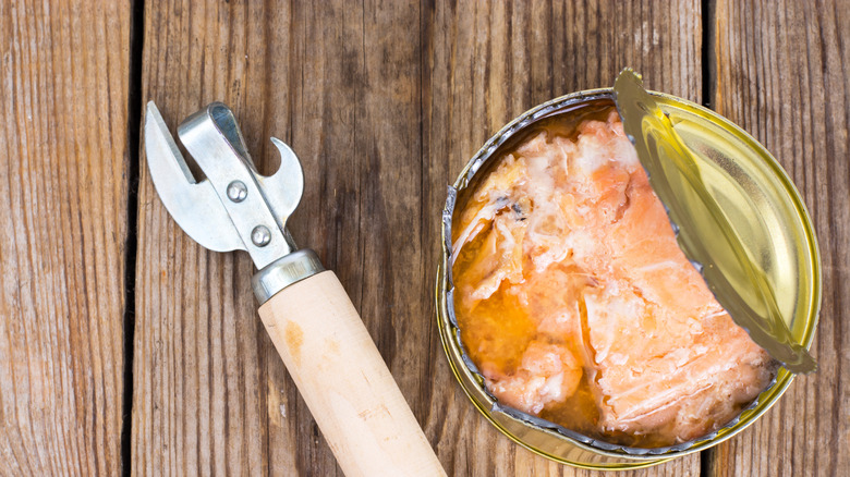 overhead view of open canned salmon beside old fashioned can opener on wooden table