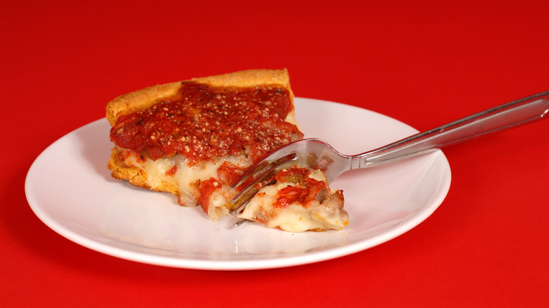 slice of deep dish pizza on white plate with fork and red background