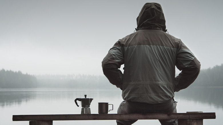 Man in coat on bench by lake with coffee