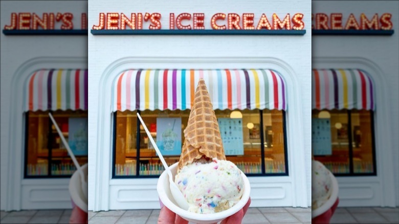 A hand holding a cup of ice cream with a cone in it in front of a Jeni's Splendid Ice Creams location