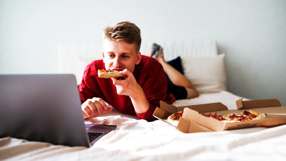 Man eating pizza on bed in front of a laptop