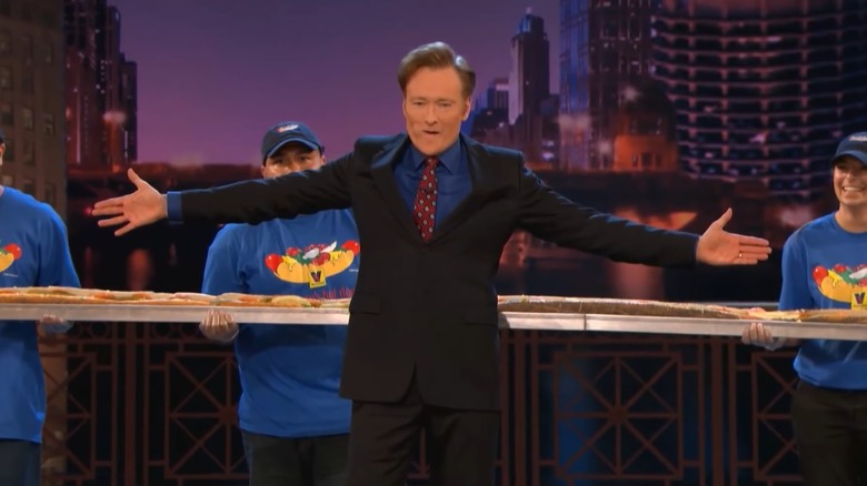 screenshot of Conan O'Brien standing in front of Vienna Beef employees holding a 40 foot long Chicago style hot dog on the set of his former late night show