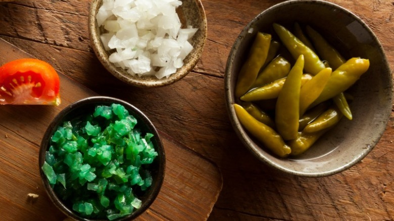 close up of small bowls of neon green relish, sport peppers, and raw diced white onion on wooden table