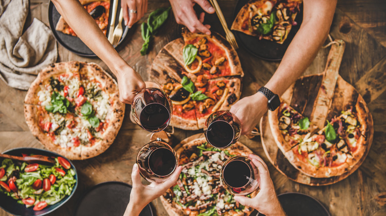 Hands clinking wine glasses around pizza