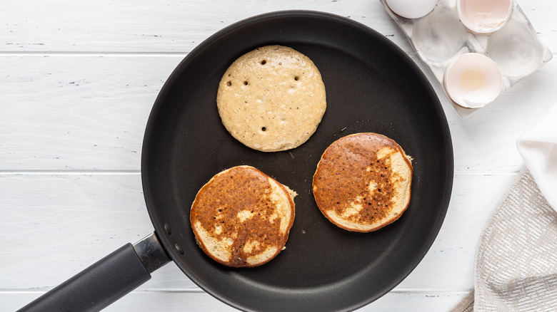 Three pancakes in a frying pan next to cracked eggs