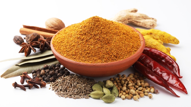 Bowl of masala powder with range of whole spices against white background