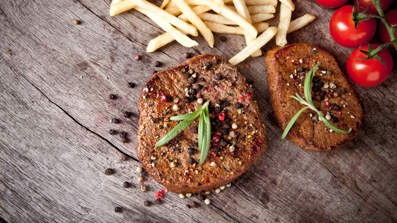 circular cuts of cooked steak with fries and tomato in background on wooden rable