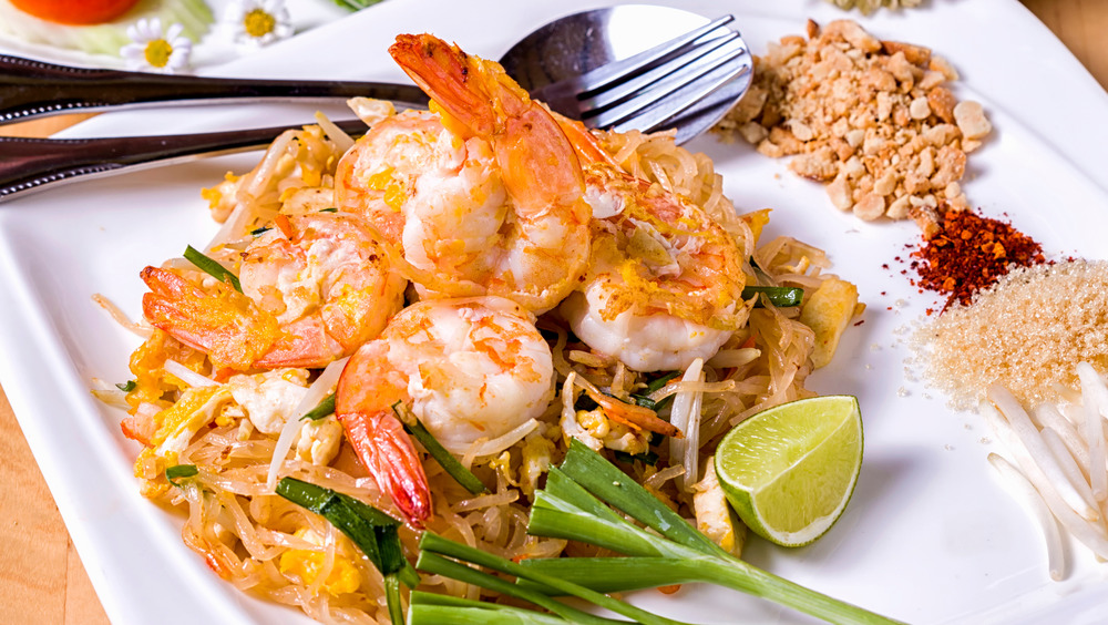 Pad Thai shrimp on white plate with fork and spoon, lime wedge, crushed peanuts, and spices