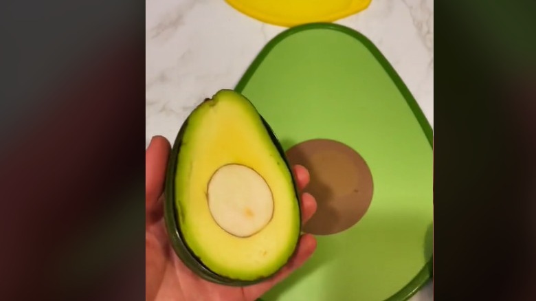 A TikTok screenshot that shows a hand holding a cut avocado, with an sliced pit, with an avocado shaped cutting board in the background