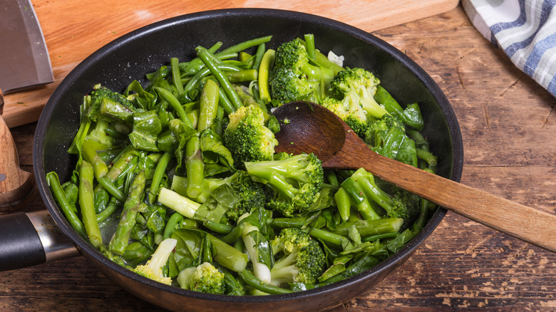 wooden spoon in frying pan crowded with cooked broccoli, green beans, asparagus, and chopped greens