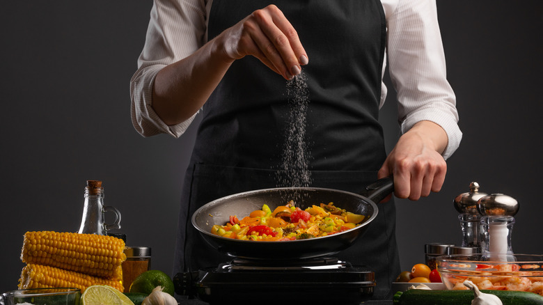 woman in black apron sprinkling salt into a frying pan of mixed vegetables