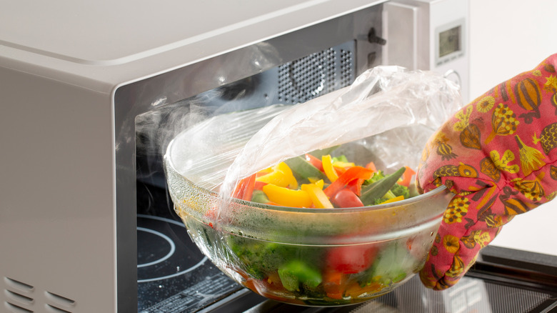 hand in oven mitt taking plastic wrap covered glass bowl of steamed vegetables out of microwave