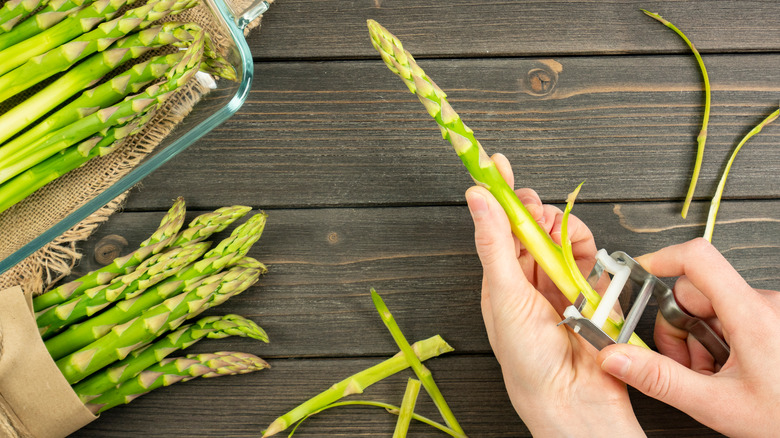 hands using vegetable peeler to trim raw asparagus stalks on wooden table