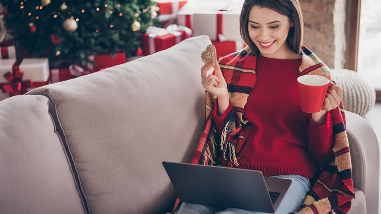 woman sitting on couch with laptop holding a gingerbread heart cookie and red mug up and smiling at her screen with Christmas tree and presents in background