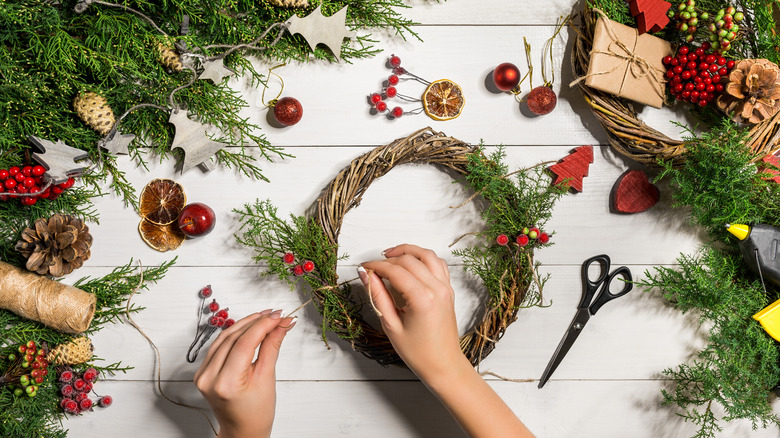 woman's hands tying evergreen sprigs and fake frosted cranberries to miniature twig wreath on white wooden table scattered with greens and Christmas decorations