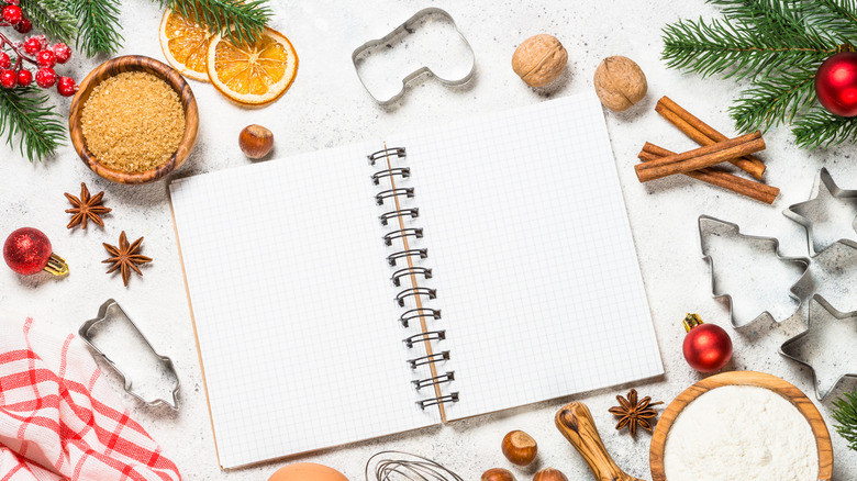 small blank spiral bound recipe book open on white table surrounded by Christmas cookie cutters, spices, sugar, and pine branches